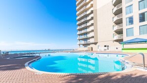 Una piscina con vista despejada al océano, rodeada por un solárium de ladrillo y un edificio de varios pisos con balcones.