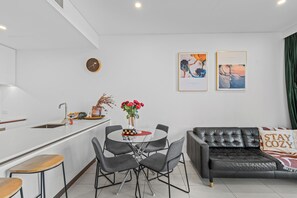 Una sala de estar moderna con un sofá de cuero negro, una mesa de comedor con sillas, una zona de cocina con un fregadero y dos obras de arte enmarcadas en la pared.