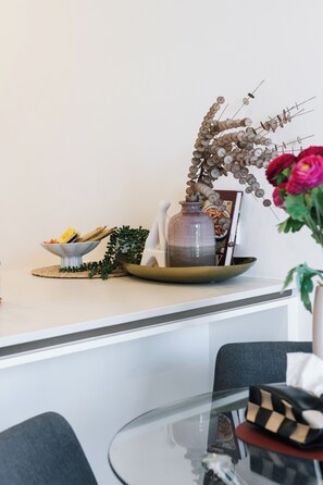 Een eetgedeelte met een tafel gedekt voor twee personen, een vaas met bloemen, en een decoratieve compositie van takken en kralen.