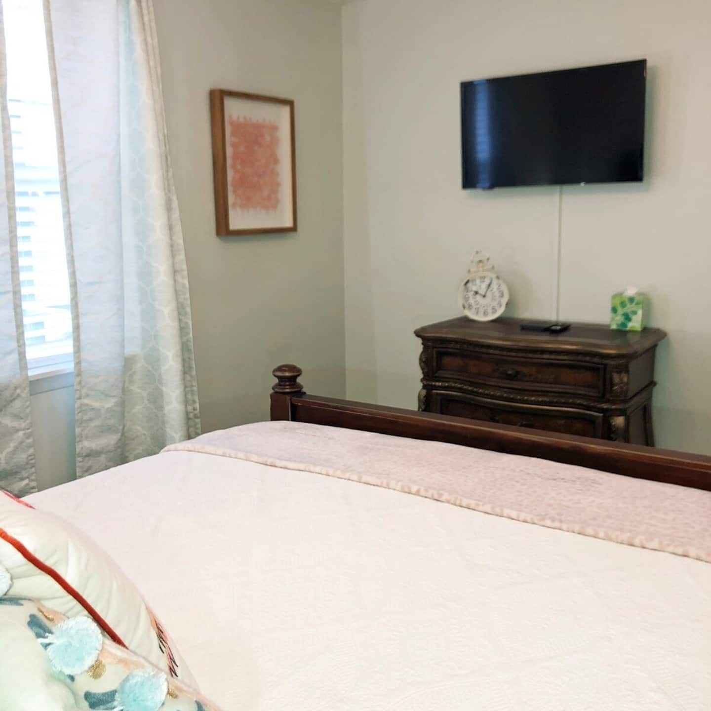 A bedroom with a bed, a dresser, a television mounted on the wall, and a window with curtains.