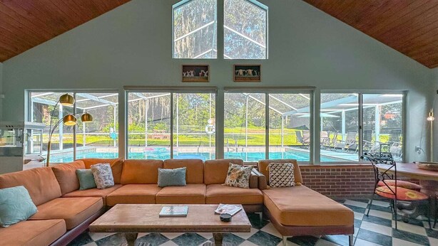 A spacious living room with a large sectional sofa, a coffee table, and a checkered floor. There are large windows offering a view of a backyard with a pool.