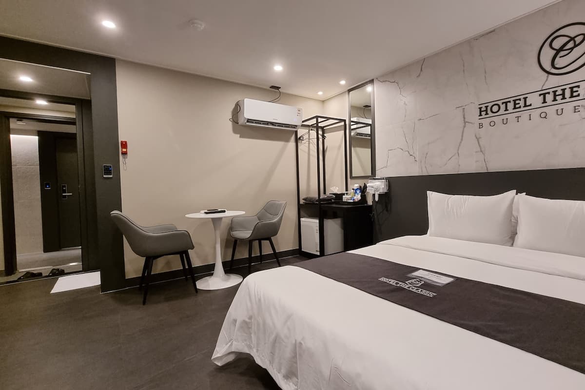 A hotel room with a bed, two chairs, a small table, a mirror, and an air conditioning unit.