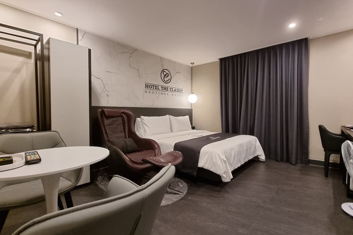 A hotel room with a bed, a desk, a chair, a table, a fan, and a wall-mounted light.