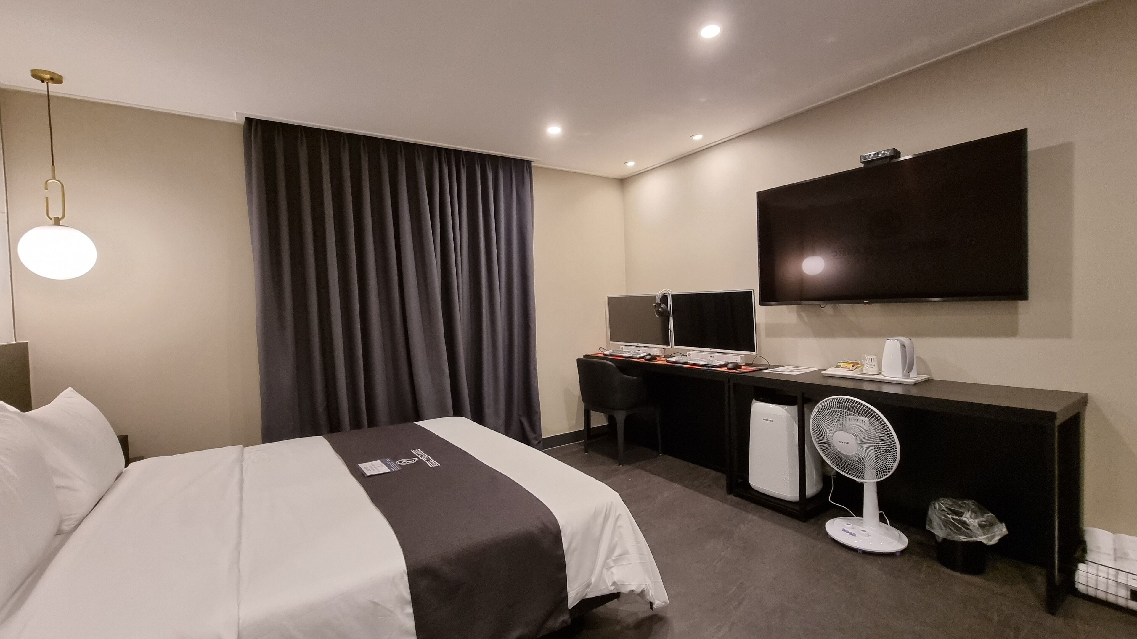 A hotel room with a bed, a desk with a computer, a flat-screen TV, a standing fan, and a large window with curtains.