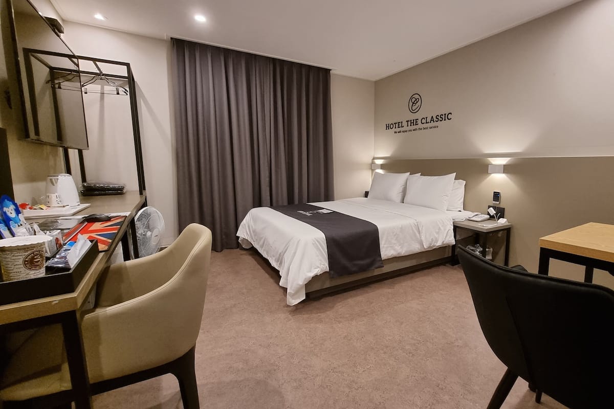 A hotel room with a bed, desk, chairs, a fan, a coffee maker, and a wall-mounted sign.