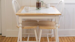 Une table à manger blanche avec deux chaises, une œuvre d’art représentant des feuilles encadrée au mur et une fenêtre donnant sur un paysage verdoyant.