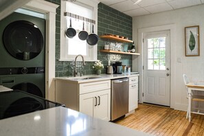 Une cuisine équipée d’un lave-vaisselle, d’un évier et d’une cuisinière. Des casseroles sont suspendues au-dessus de l’évier et une fenêtre offre une vue sur la verdure à l’extérieur.