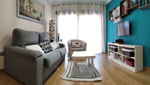 A living room with a grey sofa, a white coffee table, a patterned armchair, a painting on the wall, a TV on the wall, and a shelving unit with decorative items. - Cozy and quiet apartment (Motril)