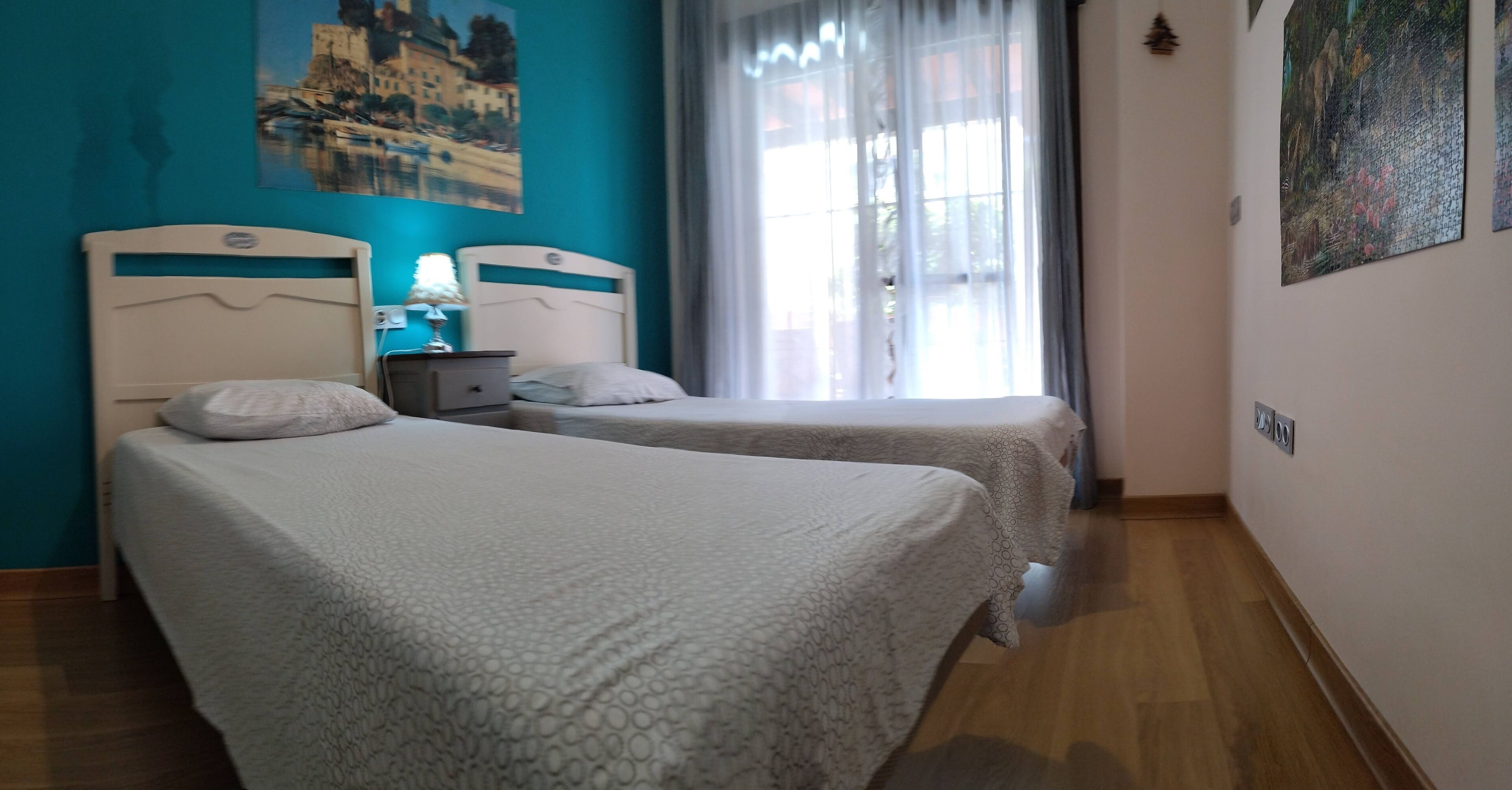 Een kamer met twee bedden, een nachtkastje met een lamp, een schilderij aan de muur en een raam met gordijnen.