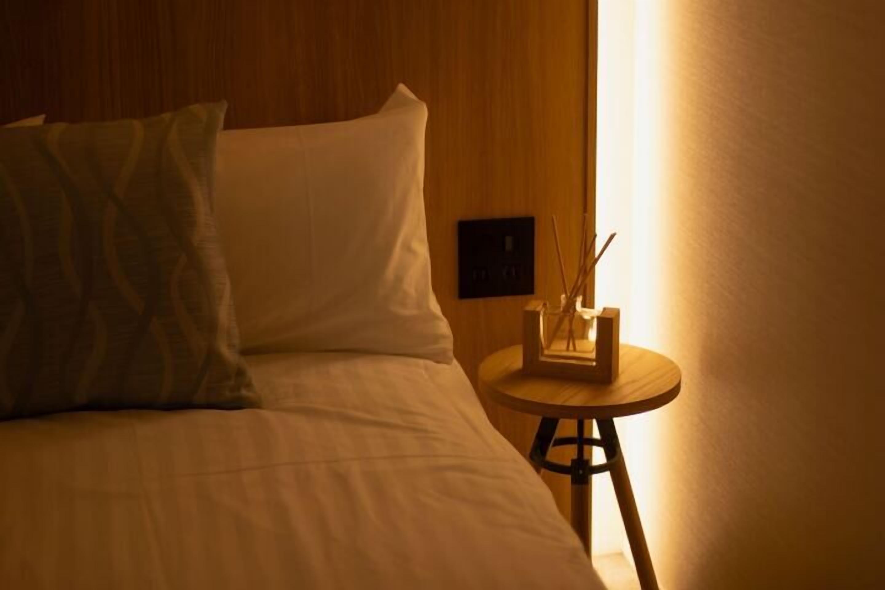 Ein Hotelzimmer mit Bett, Kissen, einem Nachttisch mit Lampe sowie einer wandmontierten Bedieneinheit.