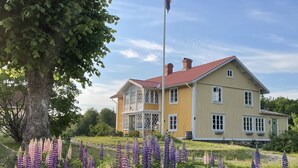 A yellow house with a red roof, surrounded by purple flowers and a large tree. - Toftahult Gård (farm) (Ryd)