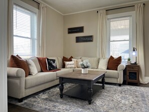 A living room with a beige sofa, a dark wooden coffee table, and a patterned rug. There are two wall-mounted signs with text, a small plant on a side table, and a lamp on a nightstand.