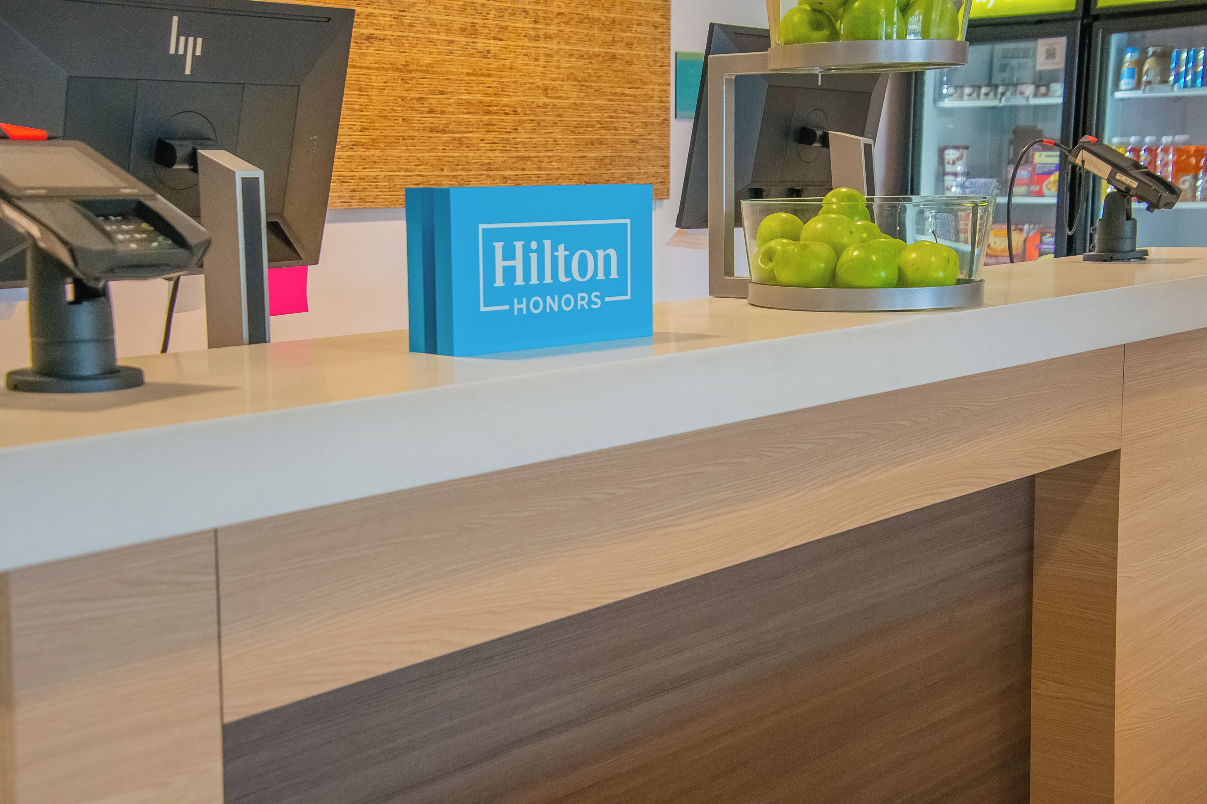 Un comptoir de réception d’hôtel avec une enseigne bleue « Hilton Honors », une caisse enregistreuse, un écran d’ordinateur, une vitrine en verre contenant des pommes vertes et, au fond, un réfrigérateur pour boissons.