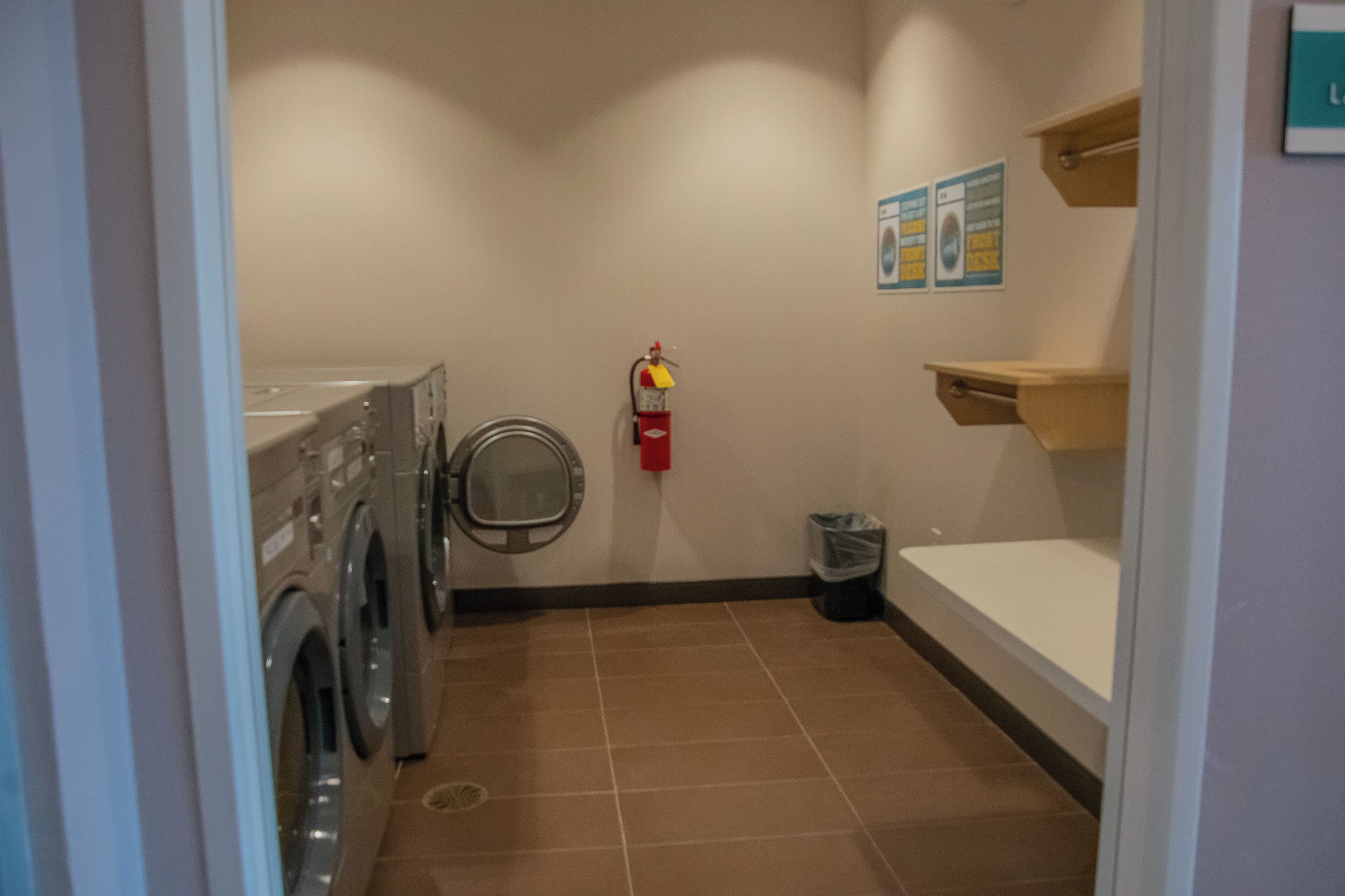 A laundry room with a washing machine, a fire extinguisher, a trash bin, and a bulletin board.
