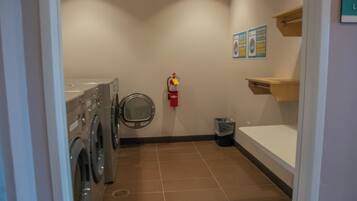 A laundry room with a washing machine, a fire extinguisher, a trash bin, and a bulletin board.