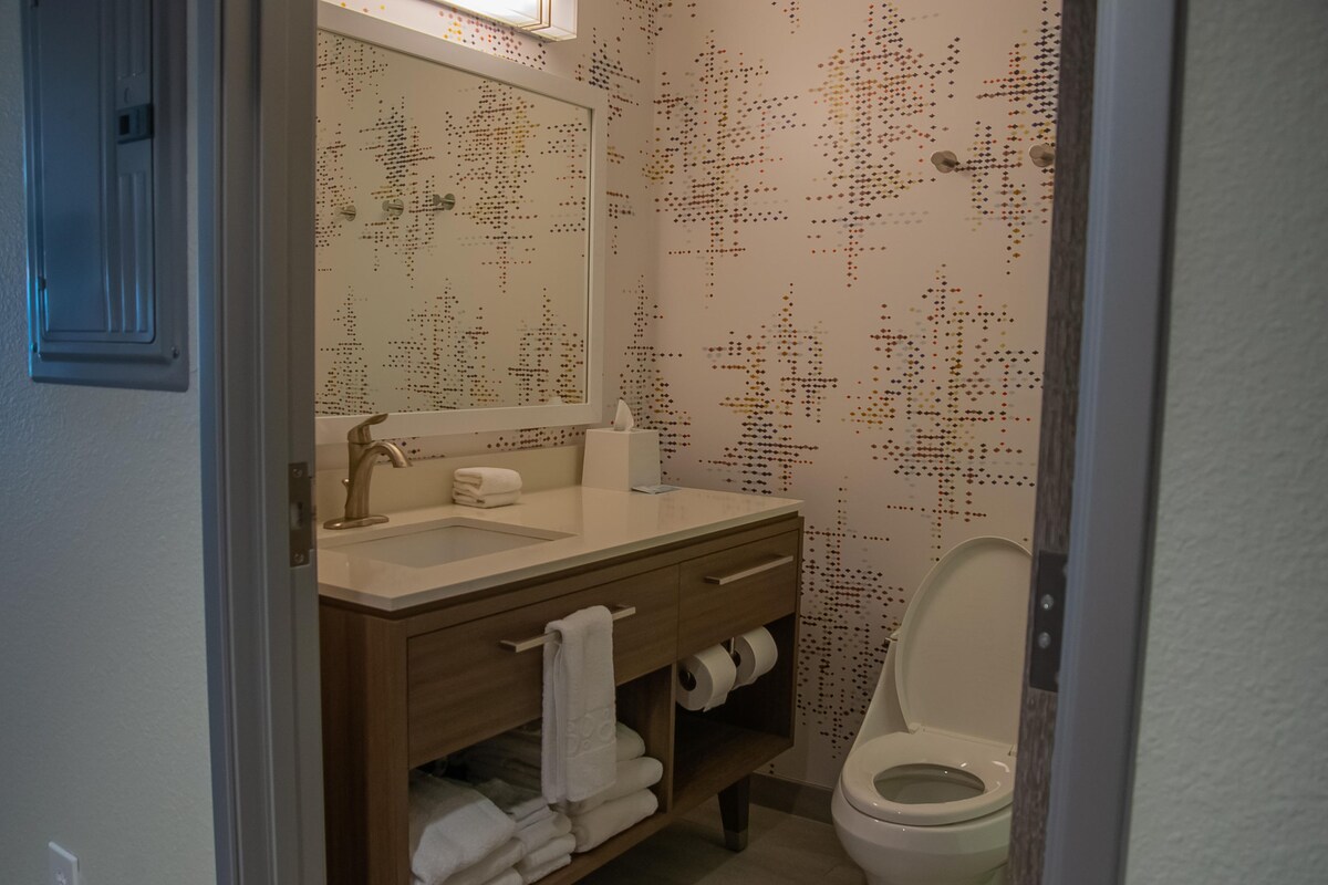 A bathroom with a wooden vanity, a sink, a mirror, a toilet, and patterned wallpaper.