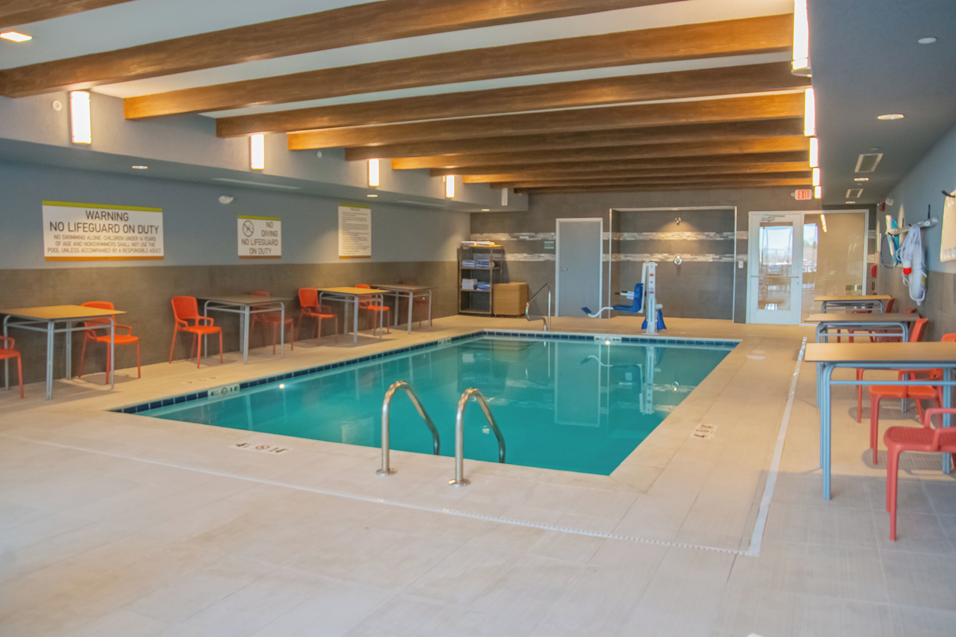 An indoor swimming pool with safety signs, wooden beams, and orange chairs.