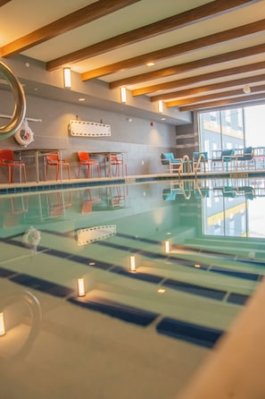 An indoor swimming pool with clear water, wooden beams on the ceiling, and orange chairs along the poolside.