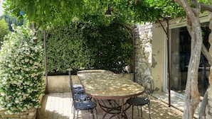 A patio with a round table and chairs, surrounded by lush greenery and a stone wall. - Gîte Les Oliviers Côté Lavande, near Vaison La Romaine  (Crestet)