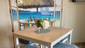 Un coin repas avec une table et des chaises, un tableau représentant une scène de plage et une petite étagère avec des plantes.