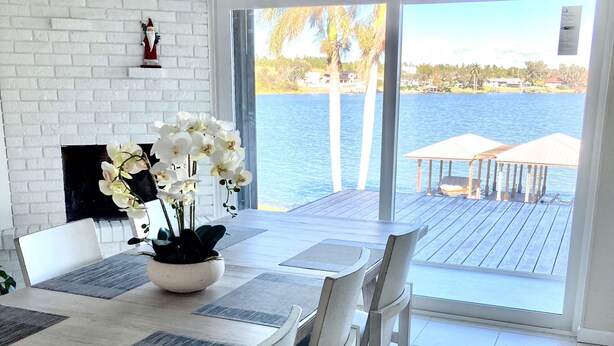 Un coin repas agrémenté d’une cheminée en briques blanches, d’une table ornée d’un vase de fleurs blanches et offrant une vue sur un lac avec un quai.