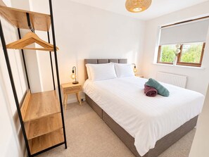 Un dormitorio con cama, lámparas de noche, un perchero de madera, un radiador y una ventana con vistas.