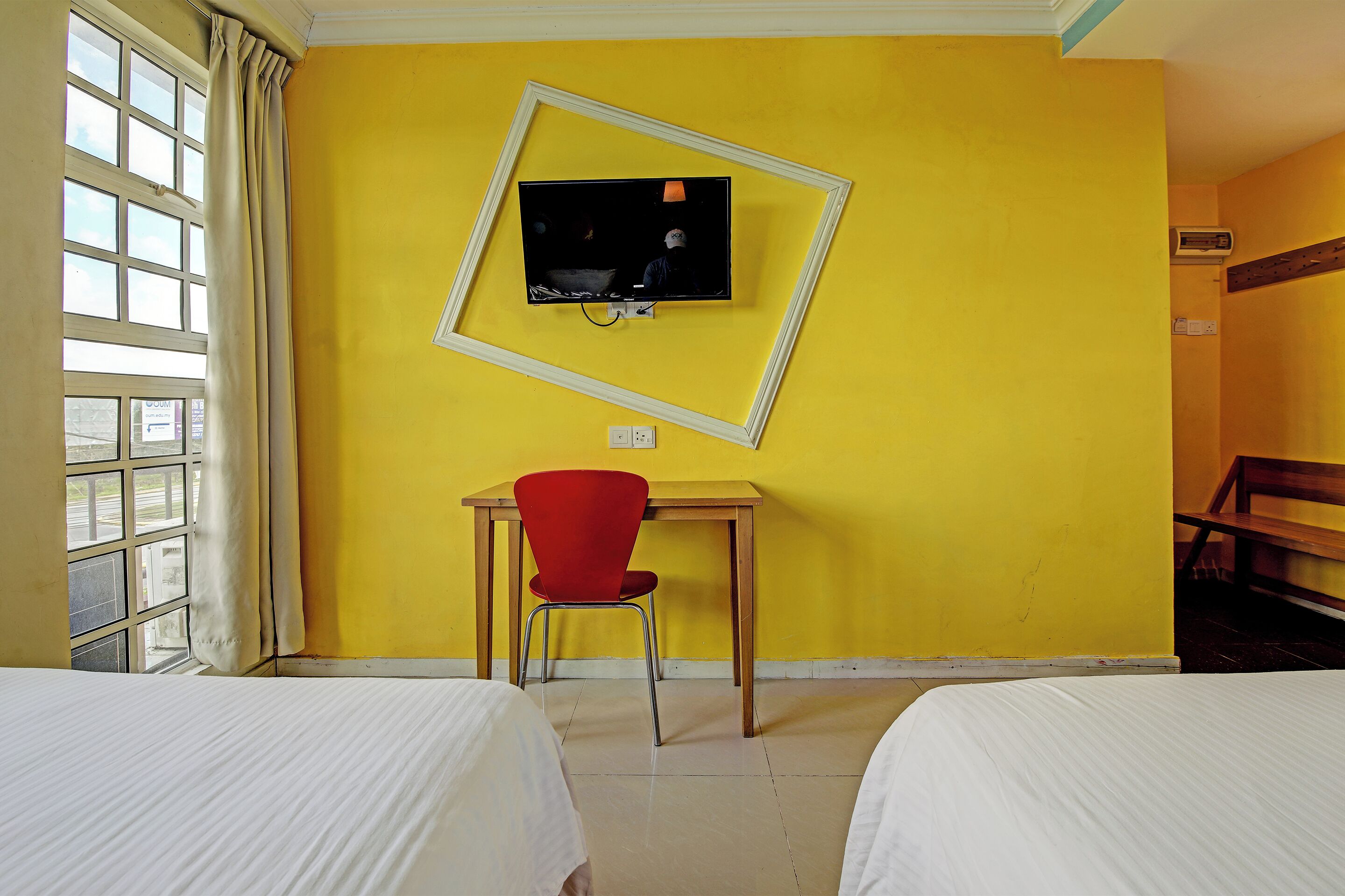 Habitación de hotel con paredes amarillas, un televisor montado en la pared, un escritorio con una silla roja y dos camas.