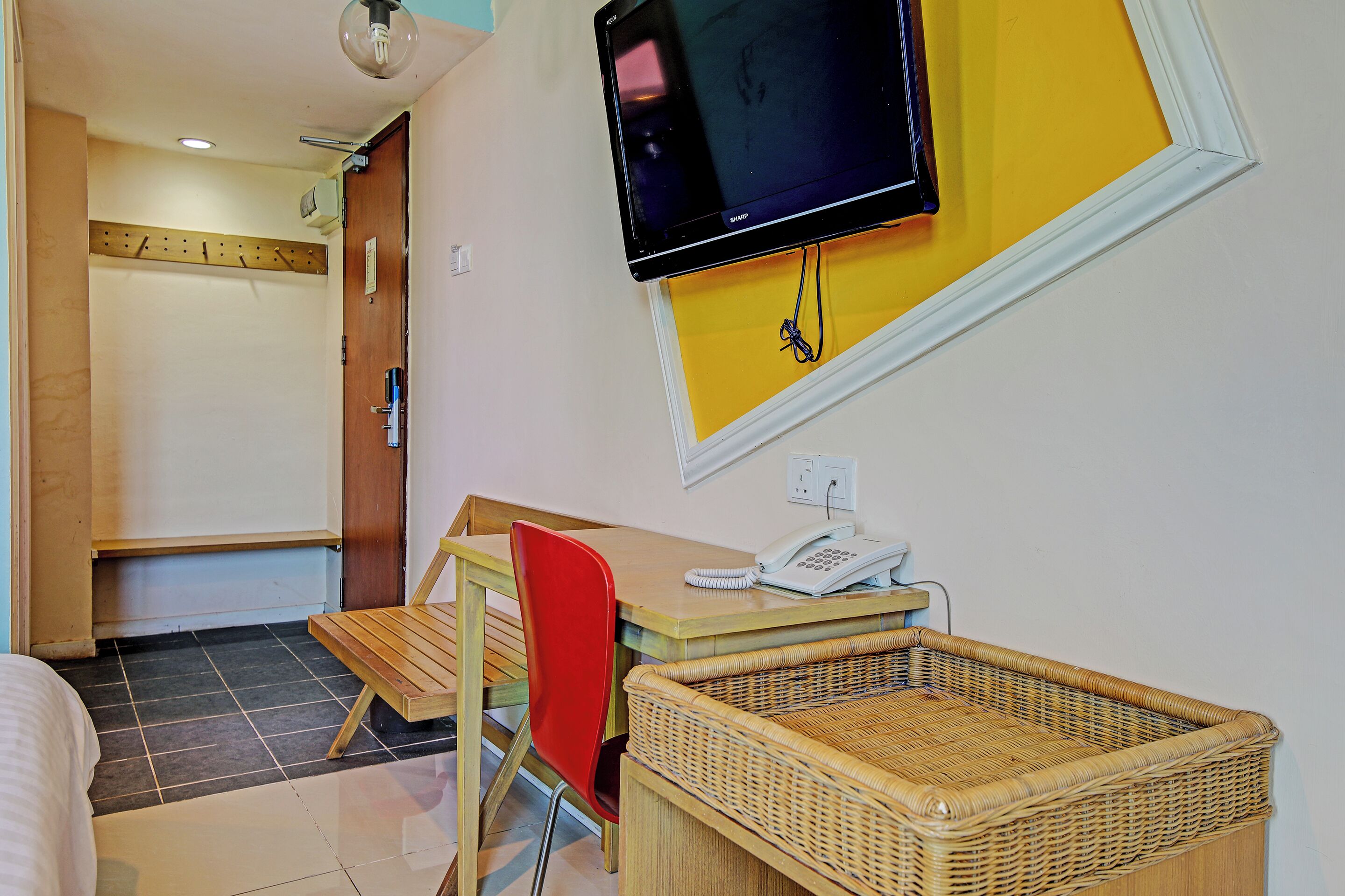A hotel room with a bed, a desk with a telephone, a red chair, a wicker basket, and a television mounted on the wall.
