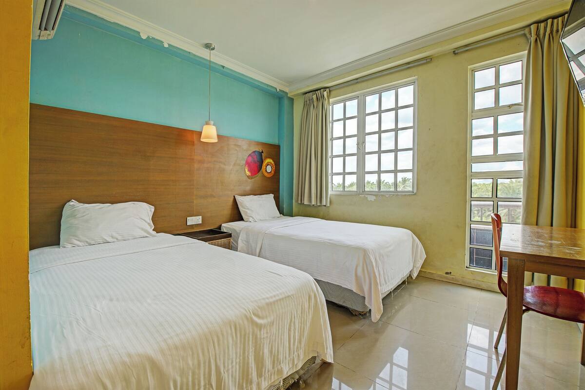 A hotel room with two beds, a wooden headboard, a table with a chair, and a window with curtains.