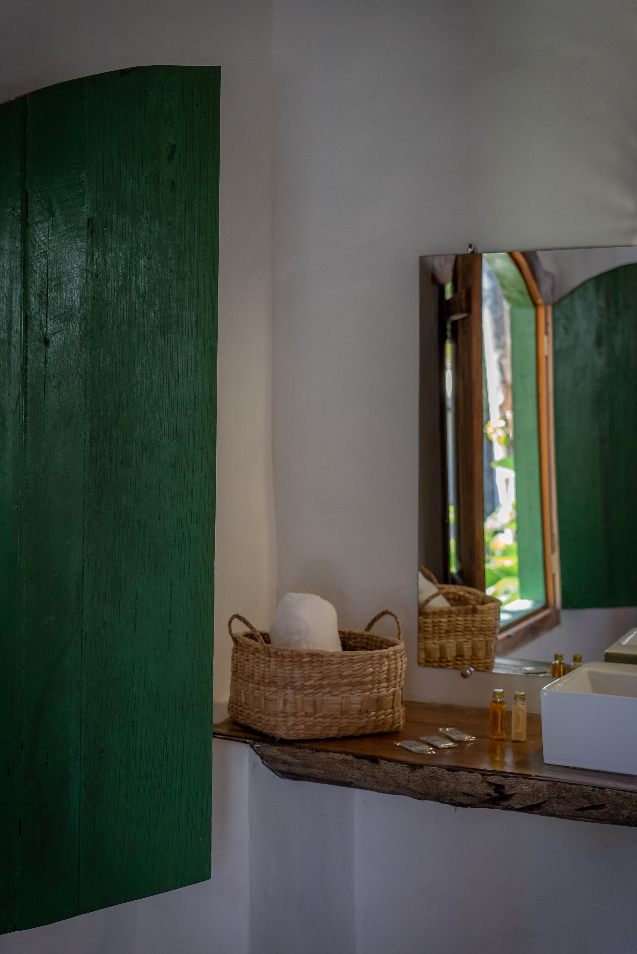 Een badkamer met een groene wand, een houten plank, een geweven mand met een handdoek en een spiegel die uitzicht geeft op een raam.