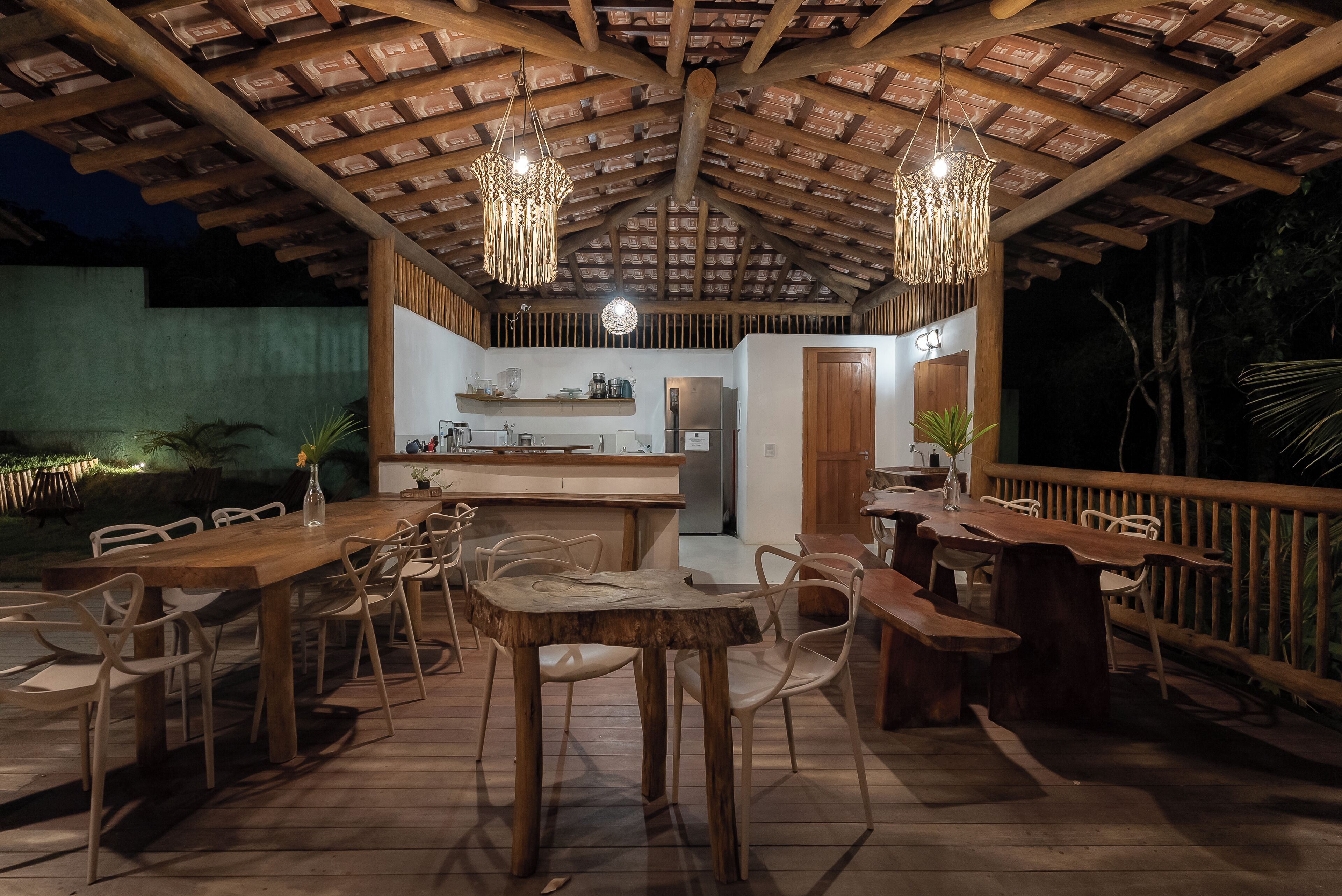 An outdoor dining area with wooden tables and chairs, a kitchen in the background, and a wooden ceiling structure.