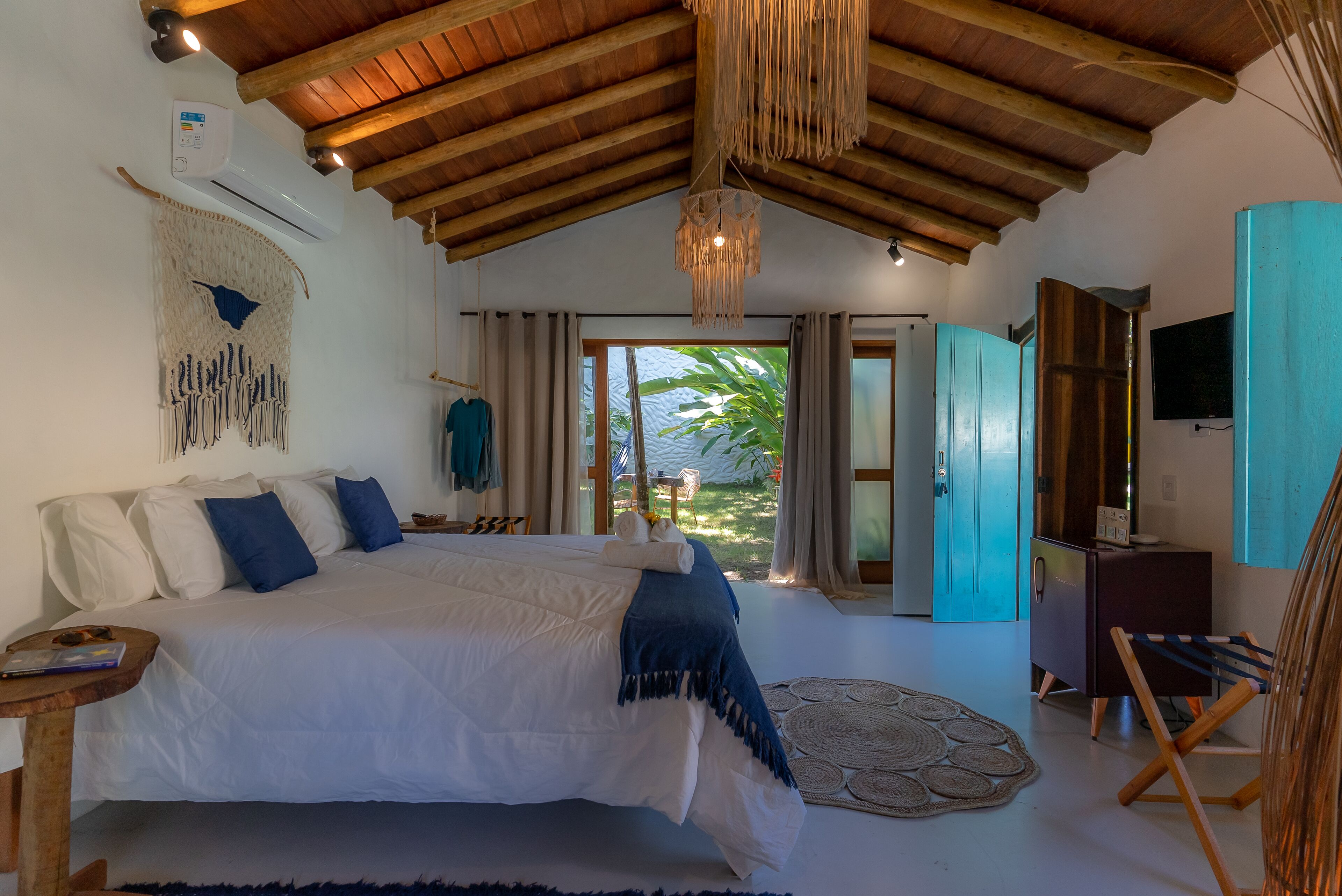Une chambre spacieuse avec un grand lit, des poutres apparentes en bois au plafond, un luminaire suspendu et une vue sur l’extérieur.