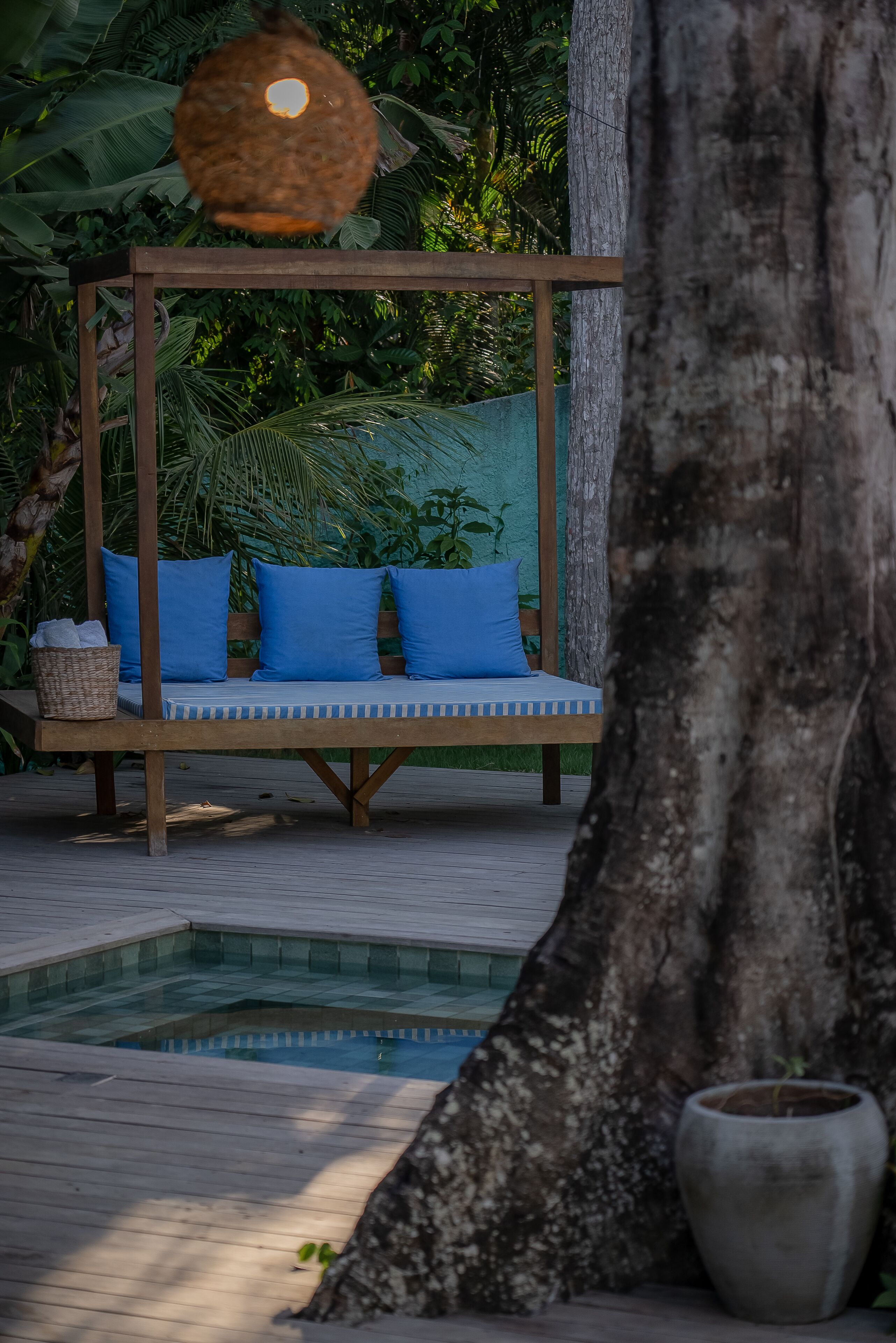 Um deck de madeira com uma cadeira suspensa, uma piscina pequena e vegetação exuberante.