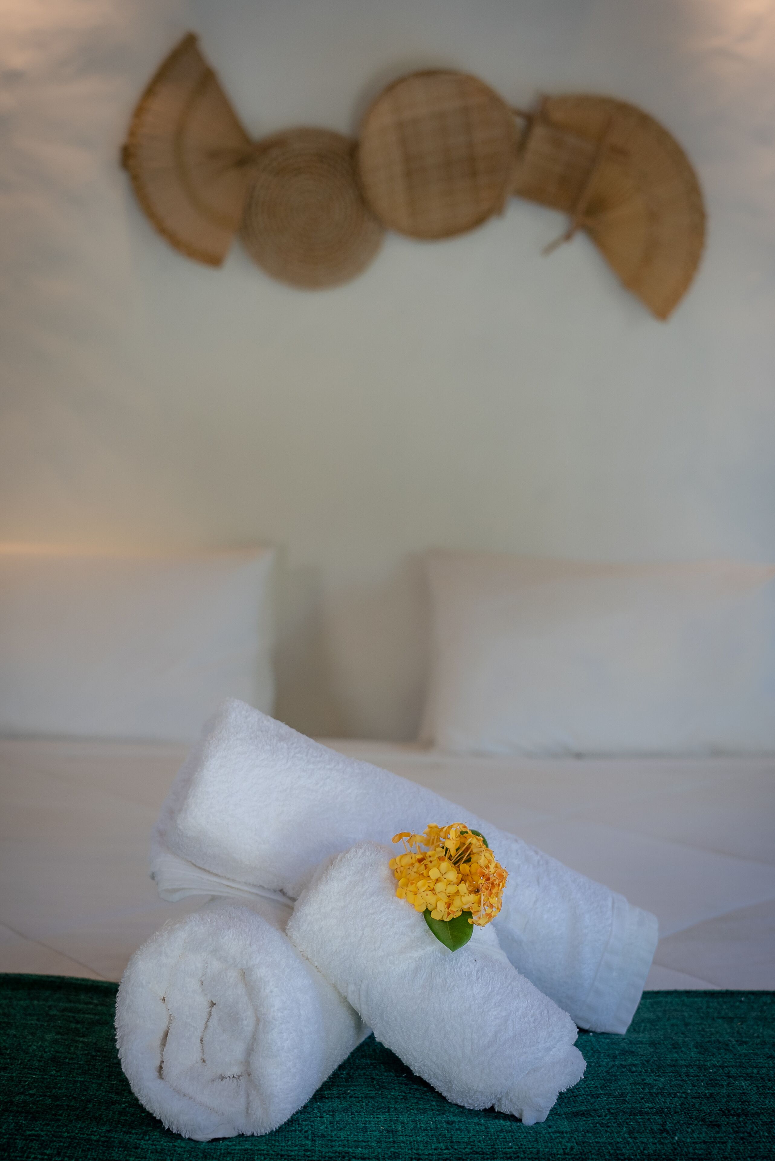 A neatly arranged set of white towels with a yellow flower on a green surface, likely a bed or a table, in a room with a white wall and a decorative wall hanging.