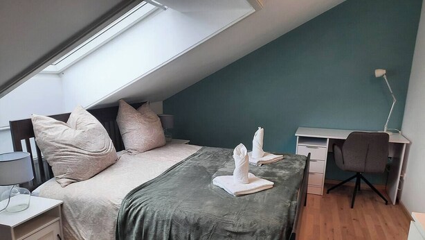 Quarto com claraboia, uma cama com manta verde, uma escrivaninha e uma cadeira.