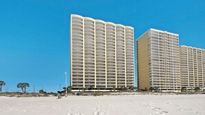 A beachfront hotel with multiple balconies, a sandy beach in the foreground, and a clear blue sky. - Gulf-Front, Sweeping Views, 2 Pools, Sleeps 6 + Free Attraction Tickets! (Panama City Beach)