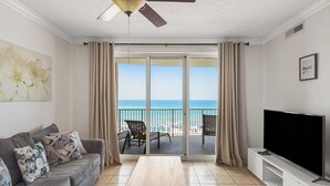 A living room with a sofa, a coffee table, a TV, and a balcony with a view of the ocean. - Gulf-Front, Sweeping Views, 2 Pools, Sleeps 6 + Free Attraction Tickets! (Panama City Beach)