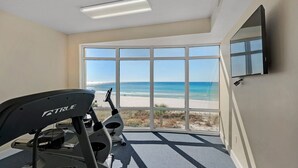 A room with a large window offering a view of the beach, a treadmill, and a flat-screen TV mounted on the wall. - Gulf-Front, Sweeping Views, 2 Pools, Sleeps 6 + Free Attraction Tickets! (Panama City Beach)