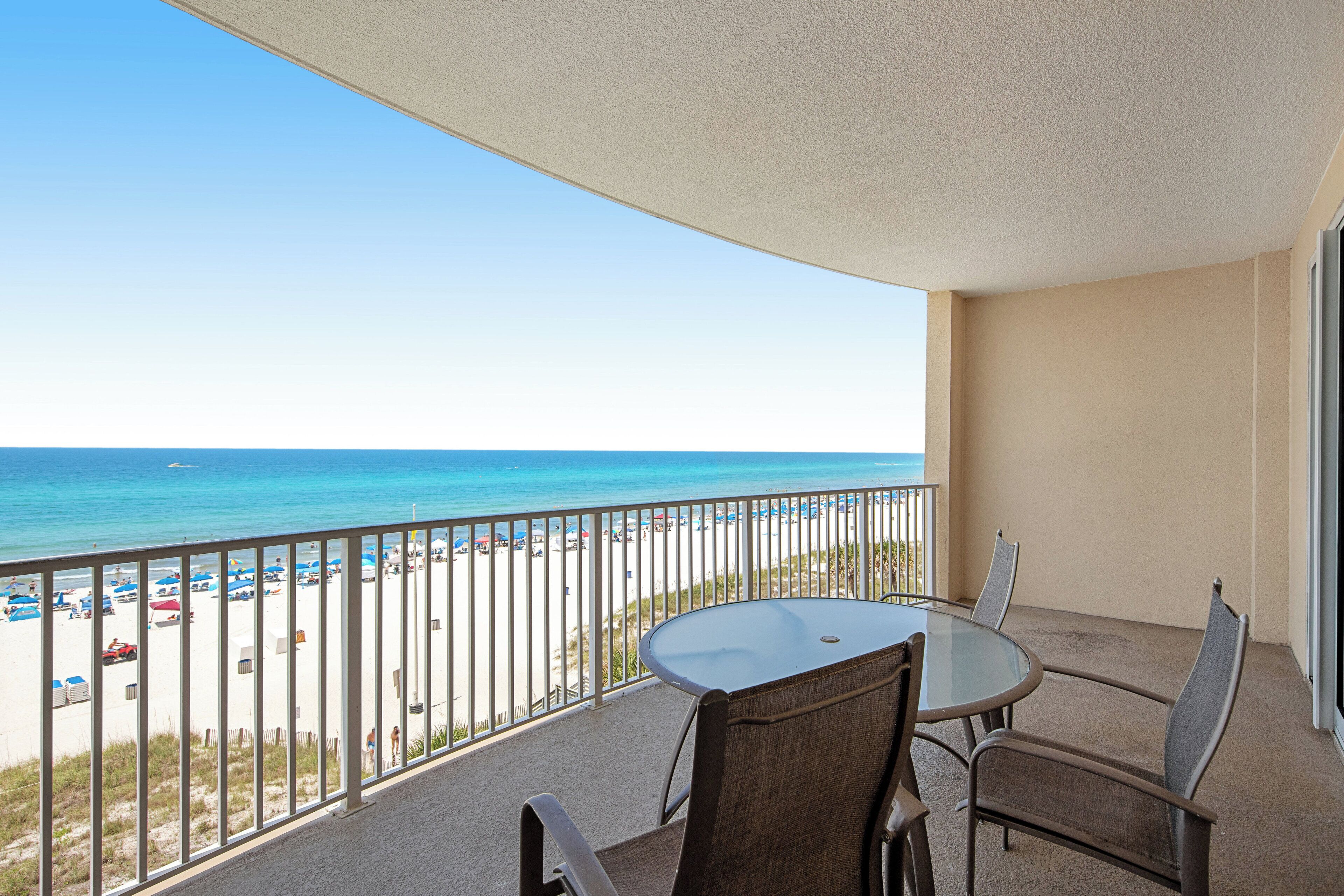A balcony with a glass table and chairs, overlooking a beach with a clear view of the ocean.