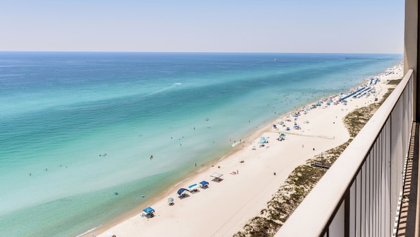 Blick auf einen Strand vom Balkon aus, mit klarem türkisblauem Wasser, einem Sandstrand und einem Steg in der Ferne.