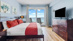 Habitación de hotel con una cama grande, televisor, cómoda de madera y balcón con vista al océano.