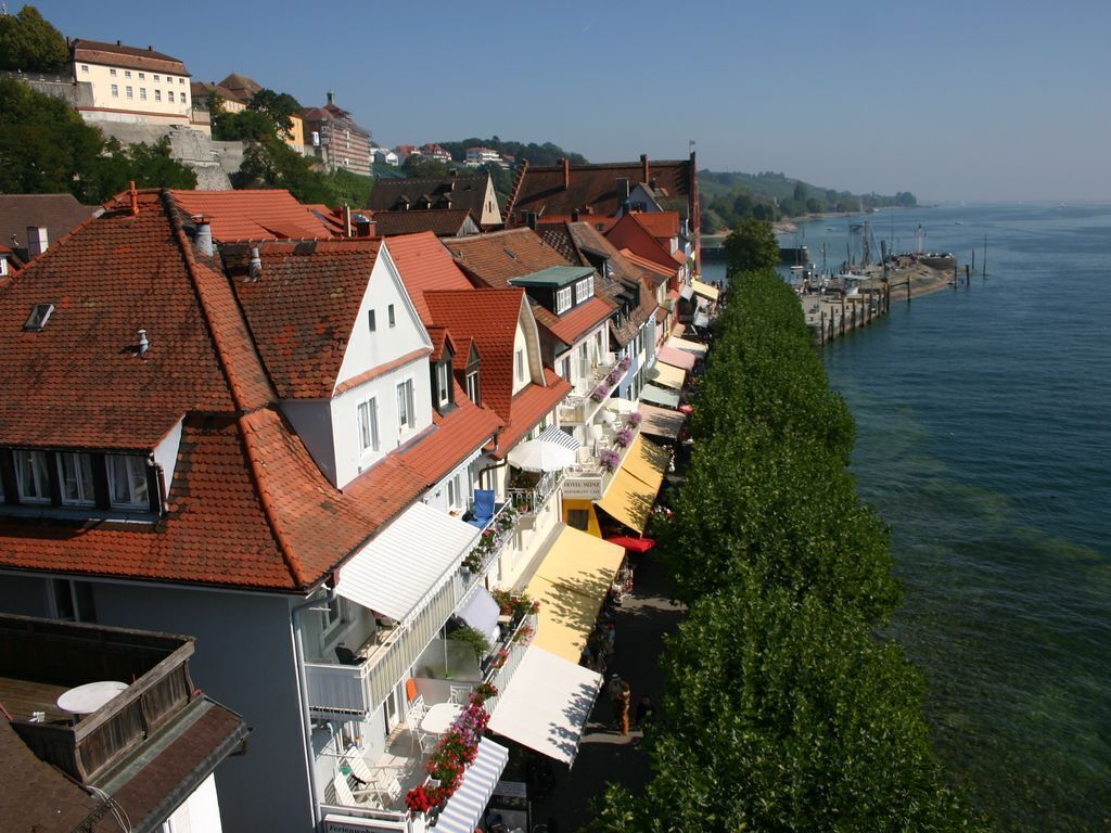 Une rangée de bâtiments aux toits recouverts de tuiles rouges longe le bord de l’eau, certains étant dotés de balcons et d’espaces de repos en plein air.
