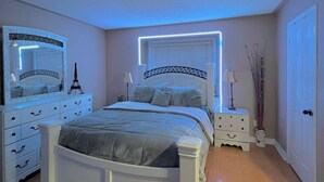 Un dormitorio con cama blanca, mesita de noche, cómoda, espejo y ventana con luces LED.