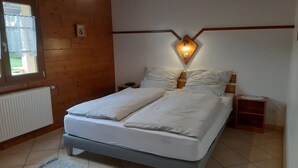 A bedroom with a bed, a nightstand, a wall-mounted light fixture, and a window with a curtain. - 2-person apartment (Soultzmatt)