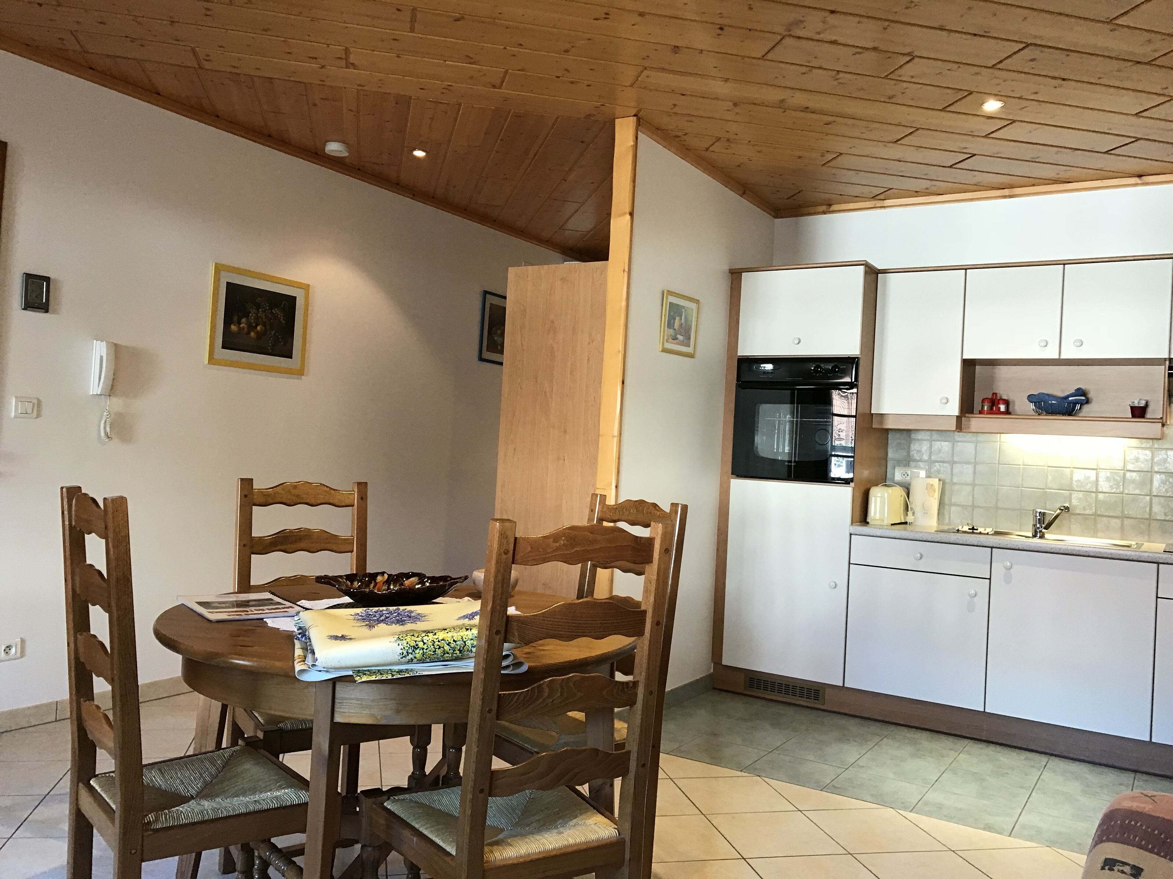 A dining area with a wooden table and chairs, a kitchen with white cabinets and a black oven, and a ceiling with wooden beams.