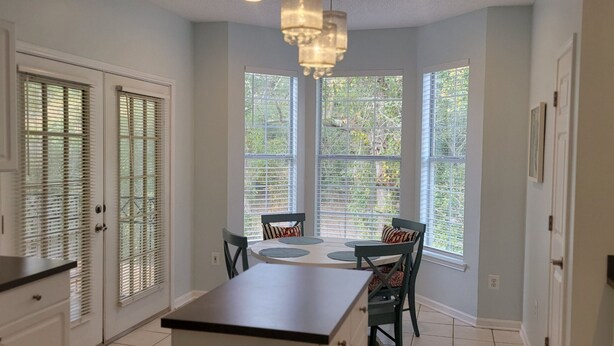 Una cocina con mesa y sillas, ventanas amplias con persianas y armarios blancos.