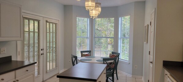 A kitchen with a dining table and chairs, large windows with blinds, and white cabinetry.
