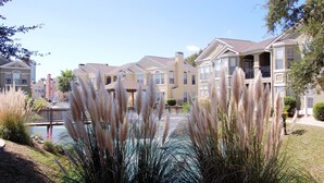 Zona residencial con varias casas de dos pisos, un pequeño puente sobre un elemento acuático y jardines bien cuidados, incluyendo hierbas altas.