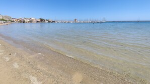 Ein Küstenstrand mit klarem Wasser, kiesigem Ufer und einem Hafen mit Booten.