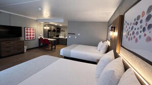 A hotel room with two beds, a TV, a desk, and a kitchenette.